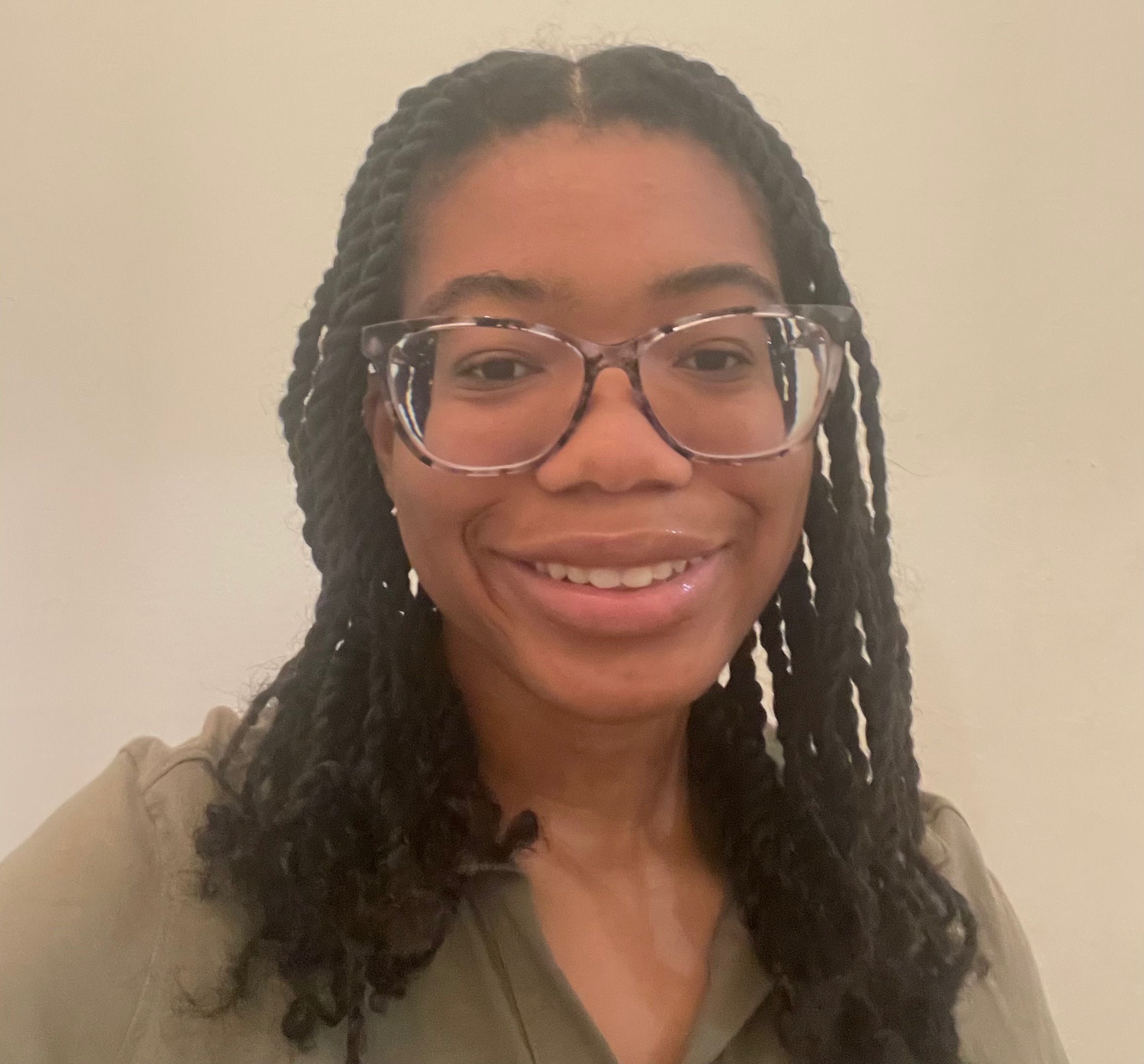 Professional picture of Ashamarie Parke, a girl of brown complexion smiling, 
        wearing a olive green top, black,light purple and black glasses,
         and there is a beige background